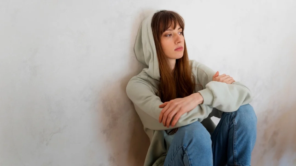 A woman wearing a hoodie is seated on the floor, appearing contemplative and relaxed in her surroundings.
