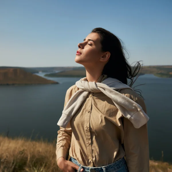 A woman standing outdoors, eyes closed, enjoying the breeze and the serenity of a natural landscape with a lake in the background.