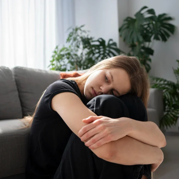 A woman sits on a couch with her arms folded, displaying a thoughtful expression in a cozy living room setting.