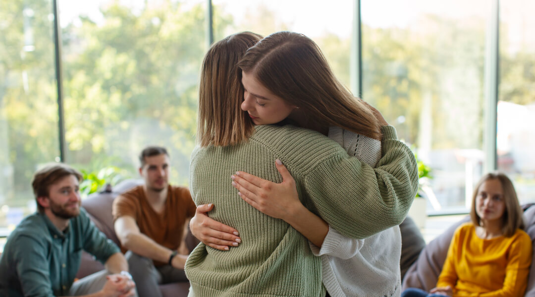 Deux femmes s'enlacent devant un groupe de personnes, exprimant une connexion chaleureuse et amicale.